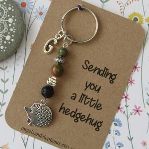May include: A brown card with the text "Sending you a little hedgehog" and a silver charm with a letter G, green and brown beads, a black lava bead, and a silver hedgehog charm attached to a silver key ring.