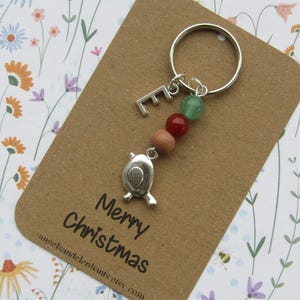 May include: A silver keyring with a bird charm, letter "E", and beads in green, red, and brown. The keyring is on a brown card with "Merry Christmas" text. The background features a floral design.