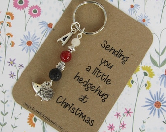 Puede incluir: Un llavero de plata con un dije de erizo, cuentas rojas, negras y blancas. El llavero está unido a una tarjeta que dice "Sending you a little hedgehog hug at Christmas".