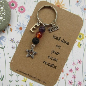 May include: A silver keyring with a star charm, a letter E charm, a 2024 charm, and three beads. The beads are amethyst, carnelian, and lava rock. The keyring is attached to a card that says "Well done on your exam results".