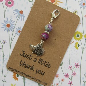 May include: A silver hedgehog charm with a lobster clasp, suspended from two beaded stones in purple and teal. The charm hangs from a brown card with the text "Just a little thank you" and the Etsy shop's URL.