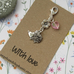 May include: A silver charm featuring a hedgehog, a pink flower, and a pink heart charm. The charm is attached to a silver lobster clasp. The charm is displayed on a brown card with the text "With love" and "angelsandelephants.etsy".