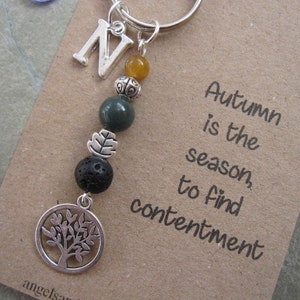 May include: A silver charm keyring with a tree charm, a letter N charm, and green and black beads. The keyring is attached to a brown card with the text "Autumn is the season to find contentment".