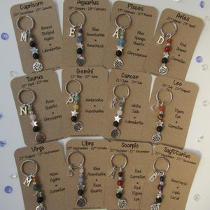 May include: Twelve keychains featuring zodiac signs and birth dates. Each keychain has a silver charm with a zodiac symbol, a letter representing the first letter of the sign, and a selection of colourful gemstones. The keychains are displayed on brown cards with the zodiac sign name and dates.