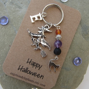 May include: A silver charm keychain featuring a witch on a broomstick, a black cat, a letter E charm, and amethyst, lava rock, and orange beads. The tag reads "Happy Halloween" and "angelsandelephants.etsy.com".