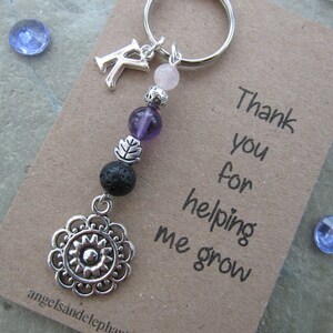 May include: A silver charm keyring with a letter 'K', a pink crystal, a purple amethyst bead, a black lava bead, and a silver flower charm. The keyring is attached to a brown card with the text 'Thank you for helping me grow'.