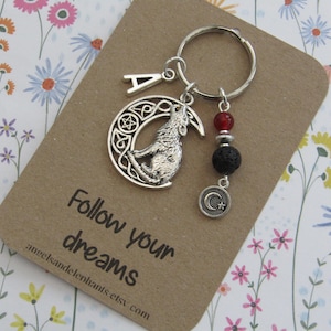 May include: A silver-coloured keyring with a howling wolf pendant inside a crescent moon design. The keyring includes a letter "A" charm, red and black beads, and a small crescent moon charm. The card reads "Follow your dreams."