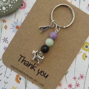May include: A silver charm keyring with a small dog charm, a letter A charm, and three beads: lavender, light green, and black lava rock. The keyring is on a brown card with the text "Thank you" and a website address.