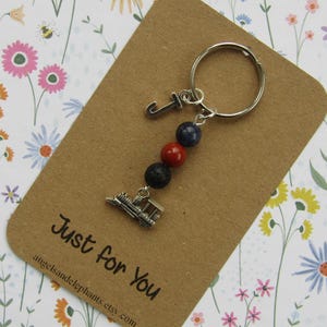 May include: A silver train charm keyring with a letter "J" charm, and three beads in red, blue, and black. The keyring is attached to a brown card with the text "Just for You".