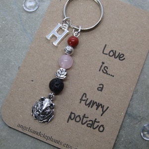 May include: A silver keyring featuring a guinea pig charm, the letter "H", and beads in red, pink, and black. The keyring is attached to a card with the text "Love is... a furry potato."
