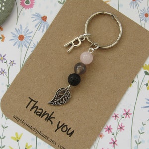 May include: A silver keyring with the letter "R" and a series of beads in pink, grey, and black. A decorative silver leaf charm hangs below the beads. The keyring is on a brown card with "thank you" printed on it.