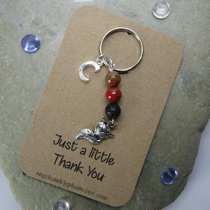 May include: A silver charm keychain with a letter C charm, red and black beads, and a silver fox charm. The keychain is attached to a brown card with the text "Just a little Thank You" and a website address.