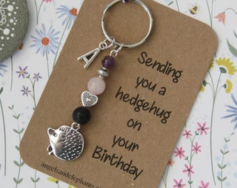 Puede incluir: Un llavero de plata con un dije de erizo, un dije de la letra A y cuentas moradas y rosadas. El llavero está unido a una tarjeta que dice "Sending you a hedgehug on your Birthday".