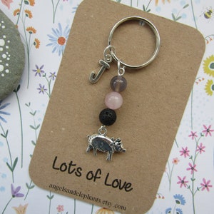 May include: A silver charm keyring with a pig charm, a letter J charm, and three beads: a dark purple bead, a light pink bead, and a black bead. The keyring is on a brown card with the text "Lots of Love" and a website address.