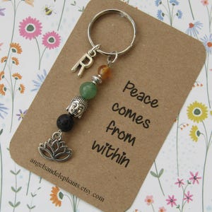 May include: A silver keyring with a green and orange beaded charm, a silver Buddha charm, a silver lotus flower charm, and a silver letter "R" charm. The keyring is attached to a brown card with the text "Peace comes from within".