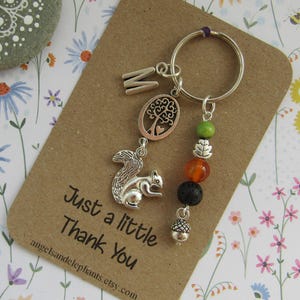May include: A silver keyring with a squirrel charm, a tree charm, a letter M charm, and a variety of beads. The keyring is attached to a brown card with the text "Just a little Thank You" and the website address "angelsandelephants.etsy.com".