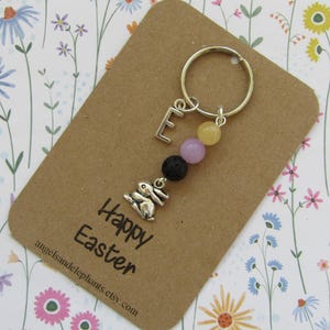 May include: A silver keyring with a bunny charm, the letter 'E', and colourful beads. The keyring is attached to a brown card with 'Happy Easter' printed on it. The background has a floral pattern.
