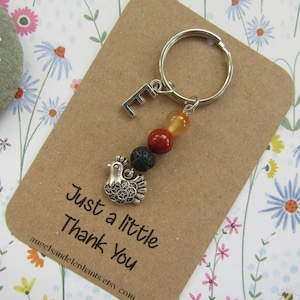 May include: A silver keyring with a small silver chicken charm, a black lava bead, a red bead, an orange bead, and a silver letter E charm. The keyring is attached to a brown card with the text "Just a little Thank You" and a website address.