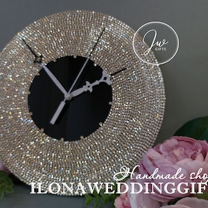 May include: A round, silver-toned clock with a black face and white hands. The clock is covered in rhinestones and has a silver-toned frame. The clock is sitting on a table with a pink flower in the background. The text "Handmade shop ILONAWEDDINGGIFTS" is visible at the bottom of the image.