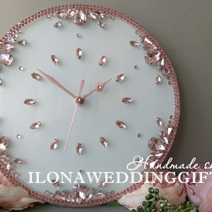 May include: A white round clock with pink rhinestones around the edge and scattered on the face. The clock hands are silver and the text "ILONA WEDDING GIFTS" is at the bottom of the clock face.
