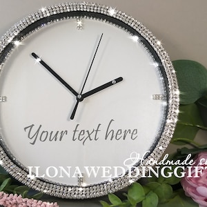 Puede incluir: Un reloj blanco con agujas negras y un borde negro. El borde está decorado con una fila de diamantes de imitación transparentes. La esfera del reloj tiene el texto "Your text here" impreso en él. El reloj está hecho por ILONA WEDDING GIFTS.