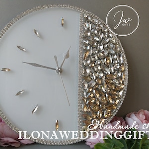 May include: A white clock with silver hands and a half-circle design of clear and gold rhinestones. The clock is surrounded by a border of clear rhinestones. The text "ILONA WEDDING GIFTS" is printed on the clock face.