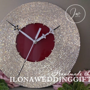 May include: A round, white clock with a red face and silver hands. The clock is covered in rhinestones and has the text "ILONA WEDDING GIFTS" at the bottom.