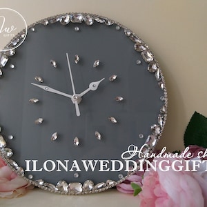 May include: A gray round clock with a white face and silver hands. The clock is decorated with rhinestones around the edge. The text "Handmade shop ILONA WEDDINGGIFTS" is visible below the clock.