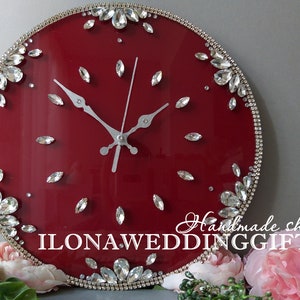 May include: A red round clock with silver hands and a silver trim. The clock face is decorated with clear rhinestones. The clock is surrounded by pink flowers. The text "Handmade shop ILONA WEDDING GIFTS" is visible at the bottom of the clock.