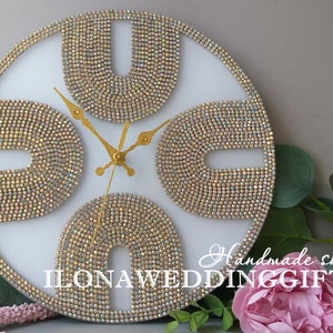 Peut inclure: Une horloge murale ronde blanche avec des aiguilles dorées et un bord doré. Le cadran de l'horloge est décoré de perles irisées dans un motif géométrique. Le texte "Handmade shop ILONAWEDDINGGIFTS" est imprimé sur le cadran de l'horloge.