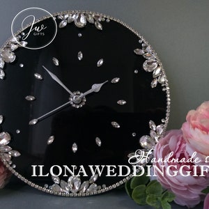 May include: A black round clock with silver hands and a silver rim. The clock face is decorated with rhinestones. The clock is surrounded by pink flowers. The text "Handmade shop ILONA WEDDING GIFTS" is visible at the bottom of the image.