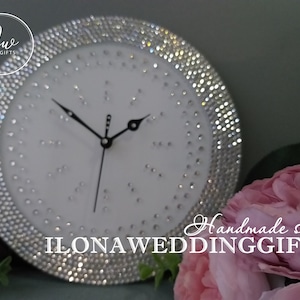 Puede incluir: Un reloj blanco con un borde de pedrería plateada. La esfera del reloj es blanca con agujas negras y un segundero negro. El reloj está sobre una mesa con una flor rosa en el fondo. El texto "Handmade shop ILONA WEDDING GIFTS" es visible en la imagen.