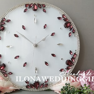 Könnte beinhalten: Eine weiße runde Uhr mit silbernen Zeigern und einem silbernen Rand. Das Zifferblatt der Uhr ist mit burgunderfarbenen Strasssteinen und klaren Strasssteinen verziert. Die Uhr ist von einem Strauß rosa Rosen und Grün umgeben. Der Text "Handmade shop ILONAWEDDINGGIFTS" ist am unteren Rand der Uhr sichtbar.