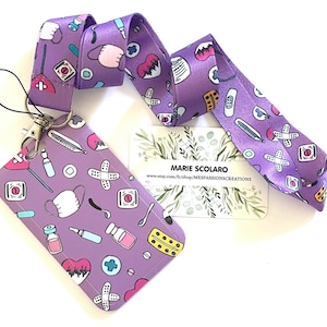 May include: A purple lanyard and matching badge holder featuring medical-themed illustrations. The lanyard and badge holder have a repeating pattern of medical symbols, including bandaids, hearts, and pills. The badge holder has a clear plastic window.