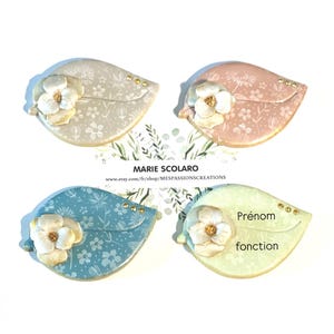 Peut inclure: Quatre décorations en forme de feuille aux couleurs pastel : beige, rose, bleu et vert clair. Chacune présente un motif floral et un accent de fleur blanche. De petits clous dorés ornent les objets. Le texte "Prénom fonction" est visible sur l'un d'eux.