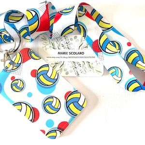May include: A white lanyard and matching card holder featuring a volleyball design with blue, yellow, and red accents. The lanyard has a metal clasp and a repeating pattern of volleyballs and colorful dots.