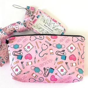 Peut inclure: Une pochette zippée rose avec une fermeture éclair noire et une sangle noire. La pochette présente un motif répété de symboles médicaux, notamment des cœurs, des stéthoscopes, des bandages et des thermomètres. Le texte "HEALTHCARD HERO" est répété sur tout le motif.