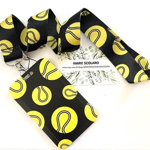 Puede incluir: Cordón y tarjetero negros con un diseño de pelota de tenis. El cordón y el tarjetero tienen un fondo negro con pelotas de tenis amarillas. El tarjetero tiene un clip plateado. El cordón tiene el texto "MARIE SCOLARO" y una dirección web.