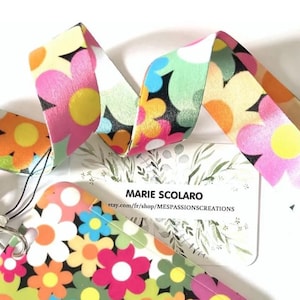 Puede incluir: Un cordón floral con un patrón repetido de flores coloridas en rosa, amarillo, azul y naranja sobre un fondo blanco. El cordón tiene un cierre de metal y una pequeña tarjeta con texto.