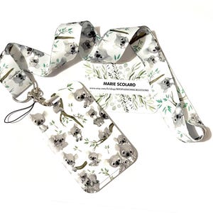 May include: White lanyard and matching cardholder featuring a repeating pattern of gray koala bears and green eucalyptus branches. The lanyard has a silver-colored clasp and a black string. The cardholder has the text "MARIE SCOLARO" and a website address.