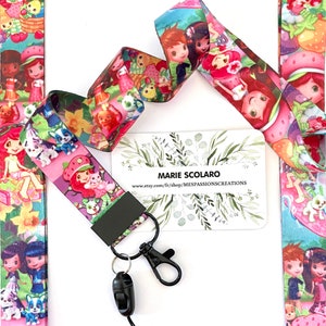 May include: A lanyard featuring a colorful cartoon design with characters, strawberries, and other elements. The lanyard has a black clasp and a black breakaway clasp. The text "MARIE SCOLARO" and a website address are visible on a white card.