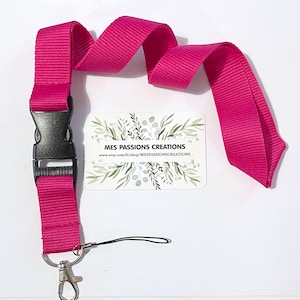 Puede incluir: Un cordón de color rosa intenso con un clip de plástico negro y un mosquetón plateado. El cordón está hecho de tejido tejido y tiene una superficie texturizada. El cordón está enrollado y los extremos son visibles. El cordón está sobre un fondo blanco. El texto en la tarjeta dice "MES PASSIONS CREATIONS" y "www.etsy.com/fr/shop/MESPASSIONSCREATIONS".