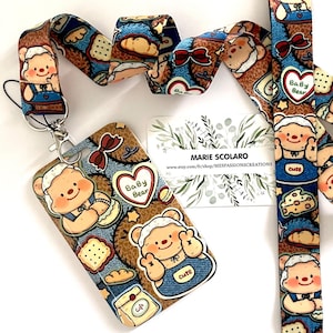 May include: A lanyard and ID holder set featuring a repeating pattern of a cute cartoon bear with a blue denim apron. The lanyard and ID holder have a brown background with images of cookies, bows, and hearts. The ID holder has the text "Baby Bear" and "cute".