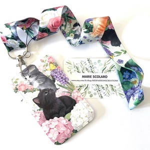 May include: A floral cat-themed lanyard with a matching card holder. The lanyard features a repeating pattern of cats and flowers. The card holder displays a black cat and a gray kitten surrounded by pink and white flowers.