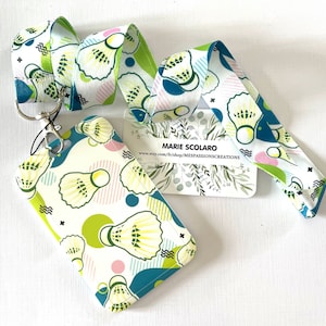 May include: A lanyard and badge holder set featuring a repeating pattern of badminton shuttlecocks in white, green, and teal. The lanyard has a silver clasp and a matching design. The badge holder has a teal border and a clear window.