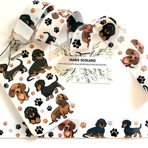 May include: White lanyard with a repeating pattern of dachshund dogs and paw prints. A matching card holder features various dachshund illustrations. The lanyard has a silver-colored clasp. The text "MARIE SCOLARO" is visible on a card.
