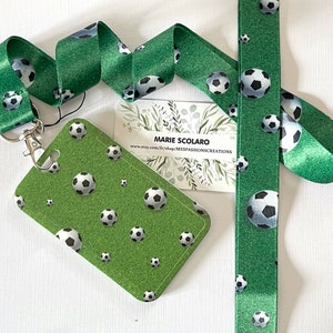 Puede incluir: Lanyard y tarjetero verdes con un patrón repetido de balones de fútbol. El lanyard tiene un cierre plateado. Una etiqueta blanca muestra el texto "MARIE SCOLARO" y una dirección web. Perfecto para aficionados al fútbol.