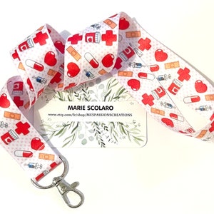 Puede incluir: Lanyard blanco con un patrón rojo, blanco y rosa. El patrón incluye corazones, vendas, píldoras y cruces. El lanyard tiene un clip de metal plateado. El texto "MARIE SCOLARO" y "www.etsy.com/fr/shop/MESPASSIONSCREATIONS" está impreso en una tarjeta blanca detrás del lanyard.