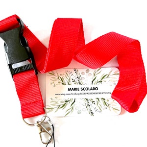 Puede incluir: Cordón rojo brillante con cierre de plástico negro y clip de metal plateado. Una tarjeta blanca con el texto "MARIE SCOLARO" y una dirección web está adjunta. La tarjeta tiene un diseño floral.