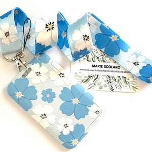 May include: A light blue lanyard and matching ID holder featuring a floral pattern of white and blue flowers. The lanyard has a metal clasp and a black cord for attaching to the ID holder. A business card is also visible.